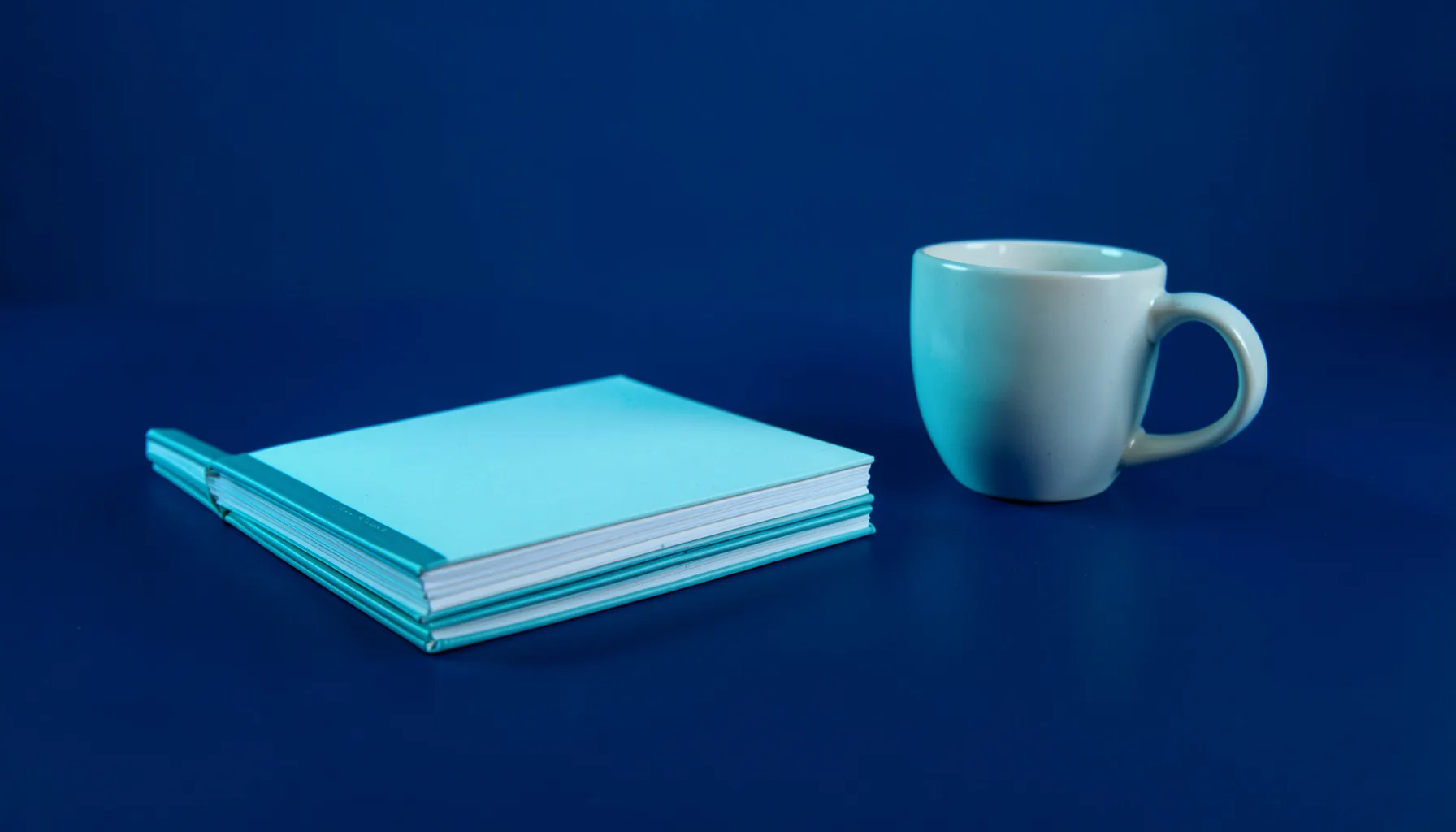 Stack of editorial notebooks and a coffee cup on a clean navy desk, lit with cyan side lighting.